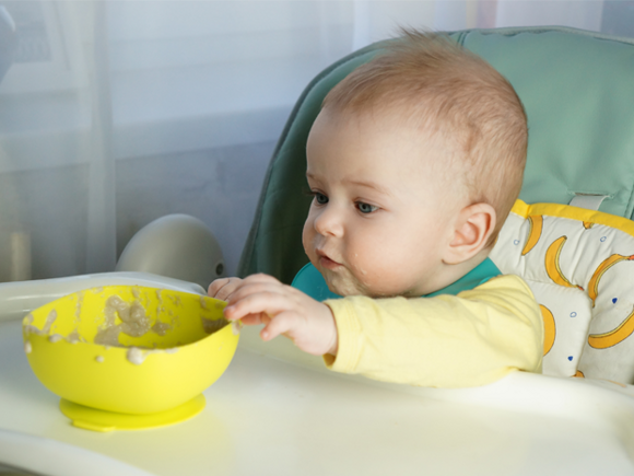 Siedzące w foteliku do karmienia niemowlę sięga po miseczkę z kaszką po 6. miesiącu