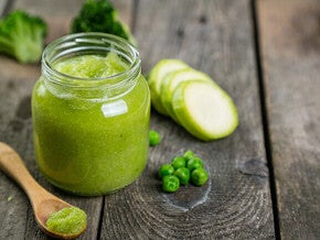 Green vegetable puree in a jar with zucchini and peas on a wooden table.