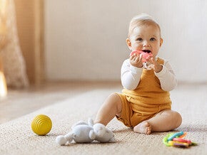 Baby in yellow overalls sitting on floor with toys nearby.