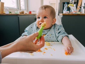 Baby in a highchair, reaching for food, kitchen background.