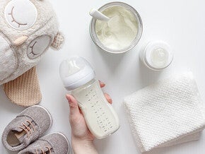 Baby essentials: bottle, formula, plush toy, shoes, and cloth on white background.