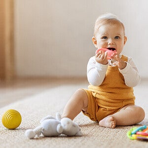 Baby in yellow overalls sitting on floor with toys nearby.