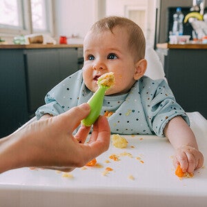 Baby in a highchair, reaching for food, kitchen background.