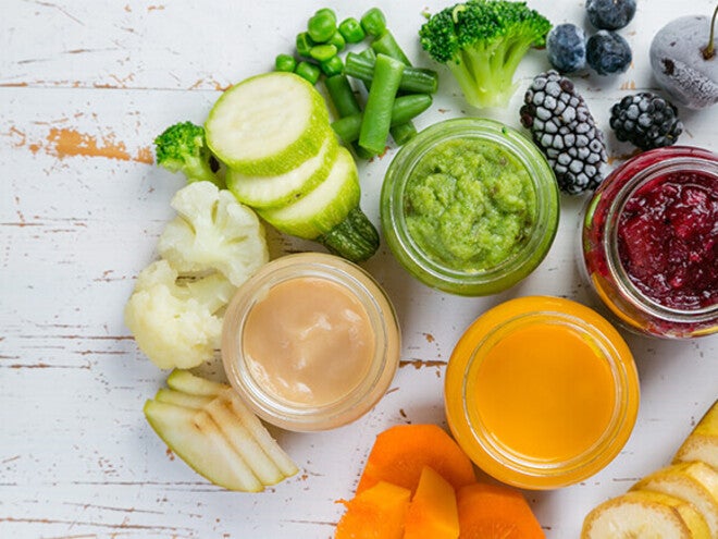 Jars of colorful purees surrounded by fresh fruits and vegetables on a rustic surface.