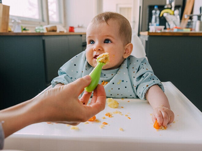 Baby in a highchair, reaching for food, kitchen background.