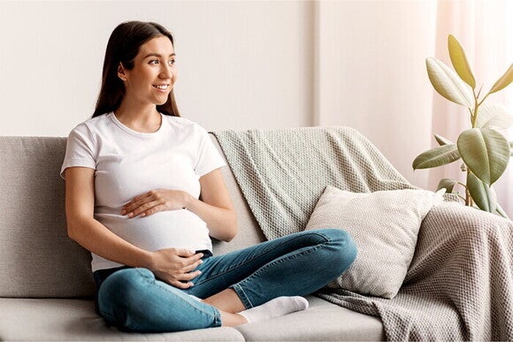 Kobieta w ciąży siedzi na kanapie, uśmiechając się i trzymając rękę na brzuchu.