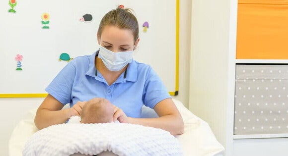 Położna środowiskowa w maseczce ochronnej badająca maluszka podczas wizyty w domu rodziny