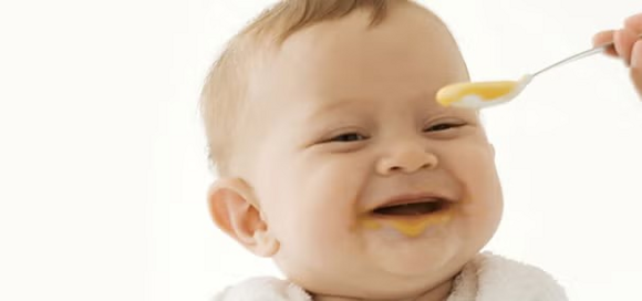 Zdjęcie uśmiechniętego dziecka w śliniaczku, karmionego łyżeczką żółtym puree, z jedzeniem na brodzie